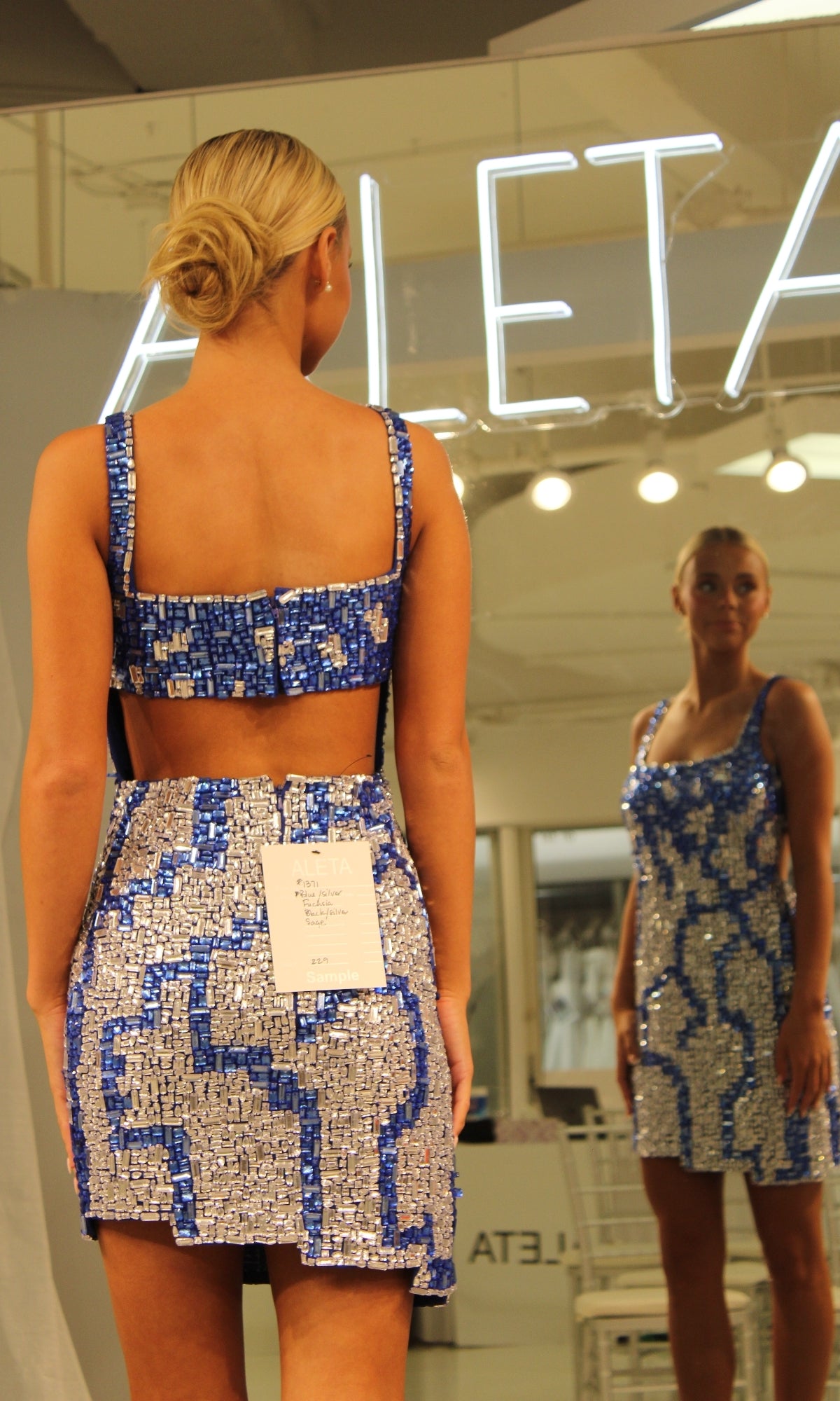 A woman in the Short Homecoming Dress 1371 by Aleta, featuring blue and silver sequins and an open back, stands before a mirror with a white tag on her skirt.